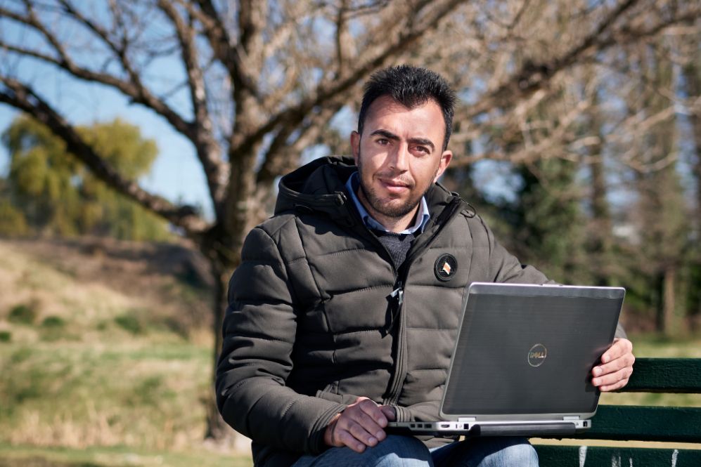 Salam Qaro sits in a park with his laptop open. He wears a black, padded jacket and jeans.
