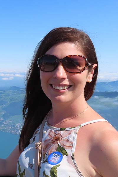 Image of Sinead Henderson smiling at the camera. Sinead is wearing sunglasses, and there is a large mountain range on the horizon. She has brown hair and is wearing a floral top.