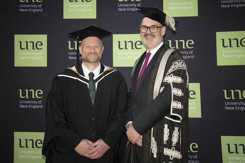 Two men in formal graduation cap and gown in front of UNE banner facing portrait to camera standing with hands resting at front
