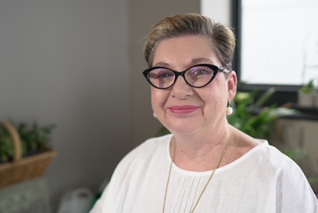 Portrait of mature woman smiling with thick black rimmed glasses and white shirt.