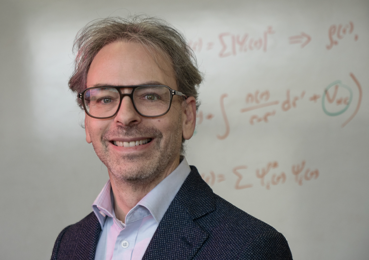 man looks at camera in front of whiteboard carrying handwritten chemical equations.