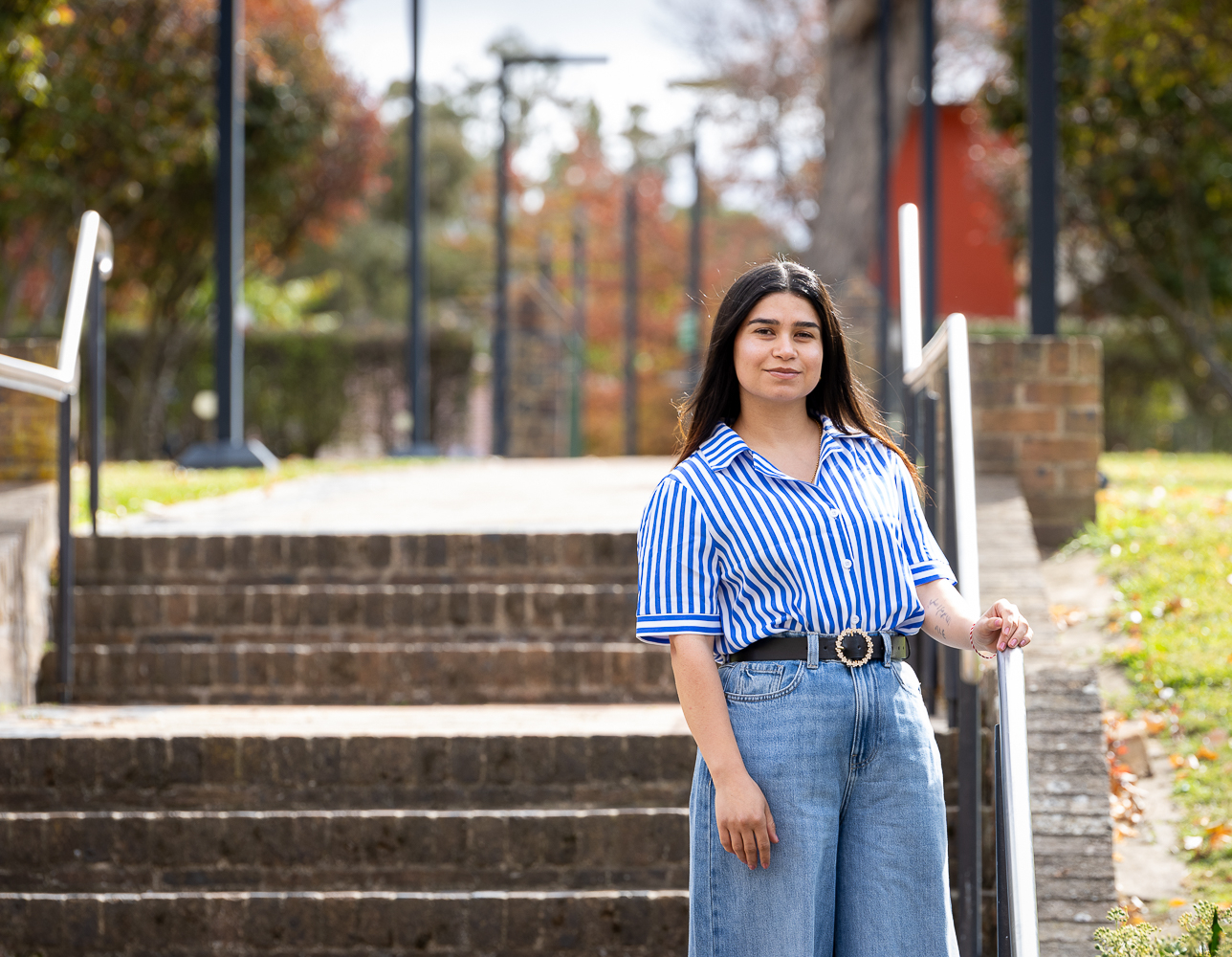 Young woman standing confidently on an outdoor staircase on a university campus in autumn. She has long dark hair and is wearing a blue‑and‑white striped short‑sleeve shirt tucked into high‑waisted wide‑leg denim jeans with a black belt. One hand rests on the metal handrail as she faces the camera with a slight smile. The background shows blurred brick steps, lamp posts, trees with green and orange leaves, and soft sunlight.