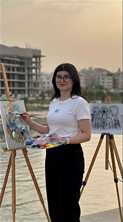 Suad Smo, a young Yazidi woman with long dark hair and glasses, stands beside a river at sunset, painting at an easel. She wears a white T‑shirt and black pants and holds a paint palette and brush. One easel displays a colourful abstract figure, while a second easel behind her shows a monochrome drawing of several outlined figures. Modern buildings and soft evening light are visible in the background.