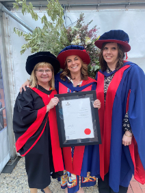 Three women dressed in doctoral graduation regalia stand together outdoors in front of a lush floral and greenery arrangement at a graduation ceremony. The woman on the left has short blonde hair and glasses, and is wearing a black doctoral gown with red trim and a black mortarboard. The woman in the centre is Olivia Tierney, smiling broadly and holding a framed Deakin University Doctor of Philosophy certificate bearing her name. She is wearing a blue and red doctoral gown with a wide-brimmed red and blue doctoral hat. The woman on the right has shoulder-length brown hair and is wearing a navy and red doctoral gown and a navy doctoral tam. All three women are smiling warmly at the camera. The setting appears to be an outdoor marquee or covered area, with brick paving visible at their feet.