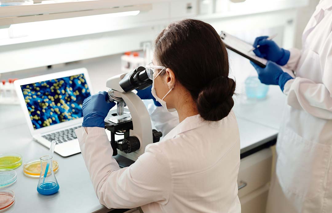 Stock image of a lady looking into a microscope. She's wearing a white lab coat.
