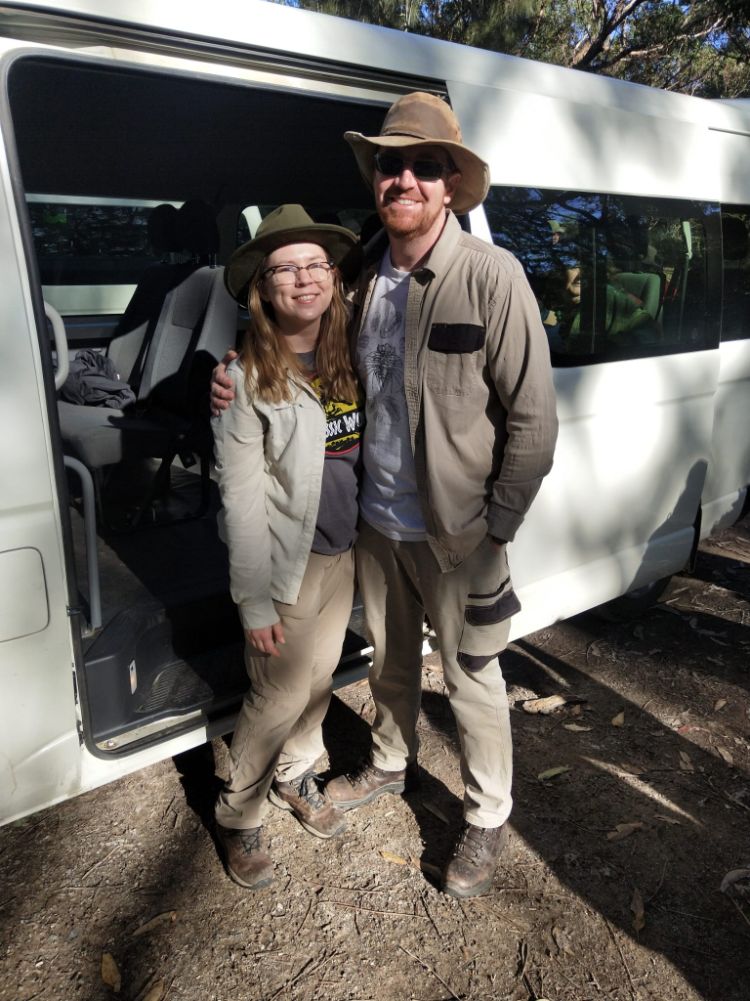 A woman and man stand at the side of a white bus parked in the bush. They are wearing hardy, khaki-coloured clothes, boots and hats.