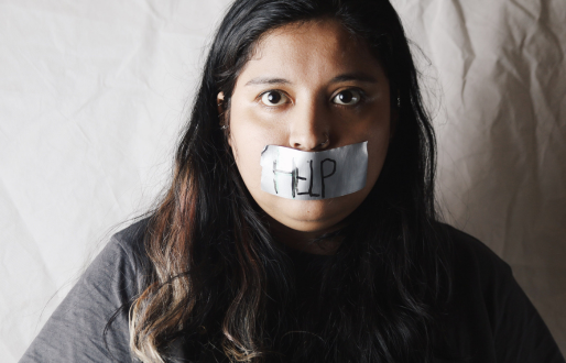 Woman of ethnic decent looks distraught at camera with her mouth tapped shut. The word 'Help' is scrawled on the tape.