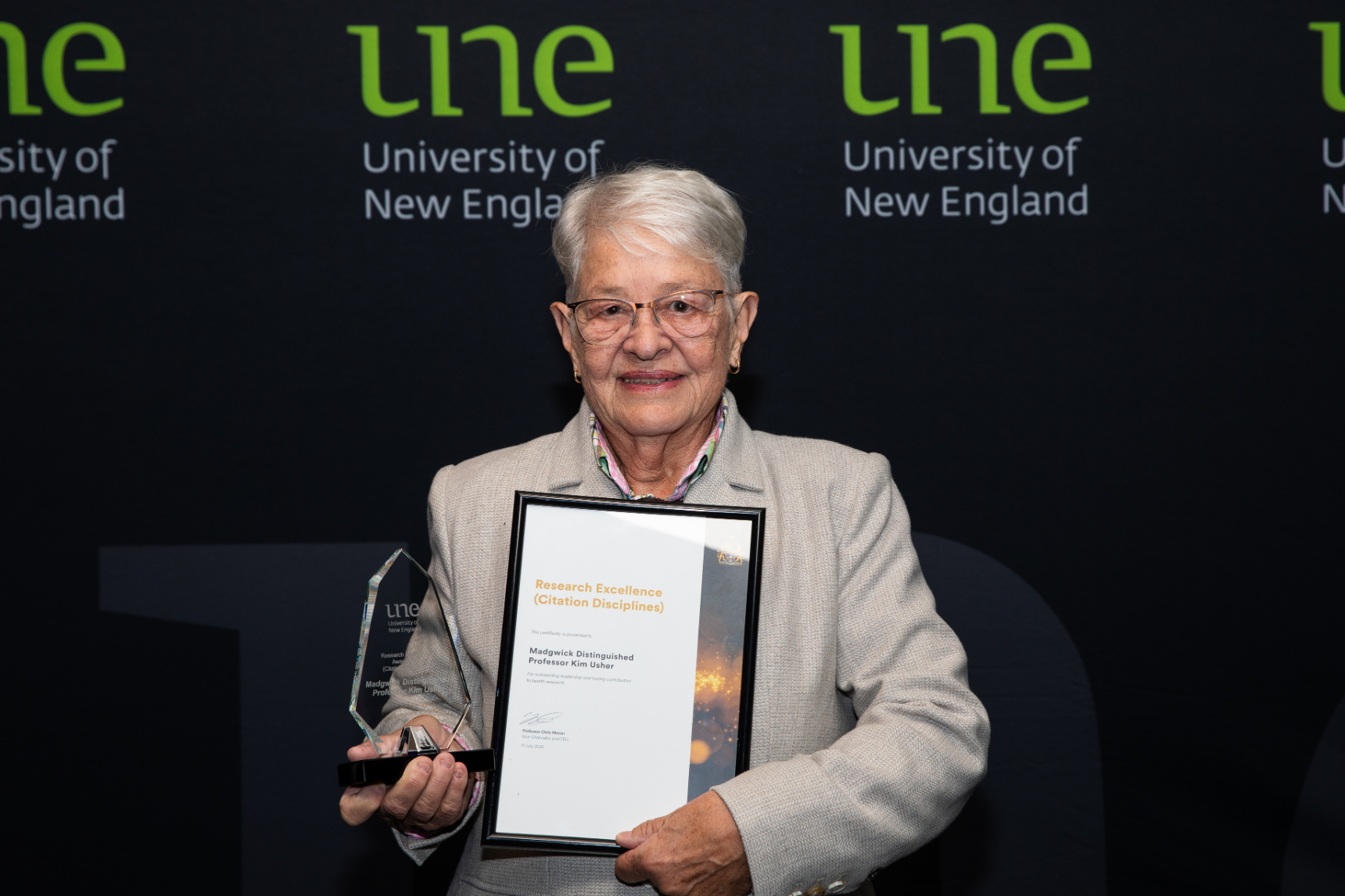 Senior female professor holding award and certificate in front of a UNE media wall at an awards night