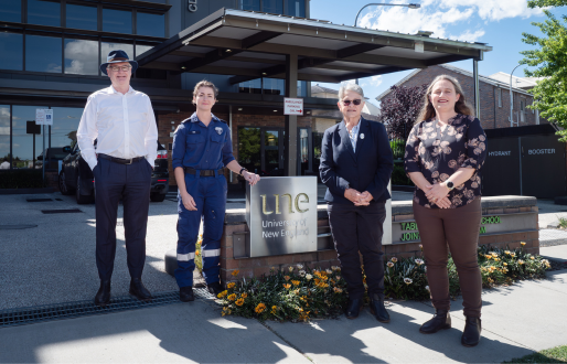 Four people stand in front of the UNE Tablelands Clinical School building at Armidale Hospital on a sunny day. From left to right: a man in a white shirt and dark trousers, a medical student in blue paramedic uniform, an older woman in a dark blazer, and a woman in a floral blouse and brown trousers. The modern glass building entrance is visible behind them with the UNE University of New England sign in the foreground.