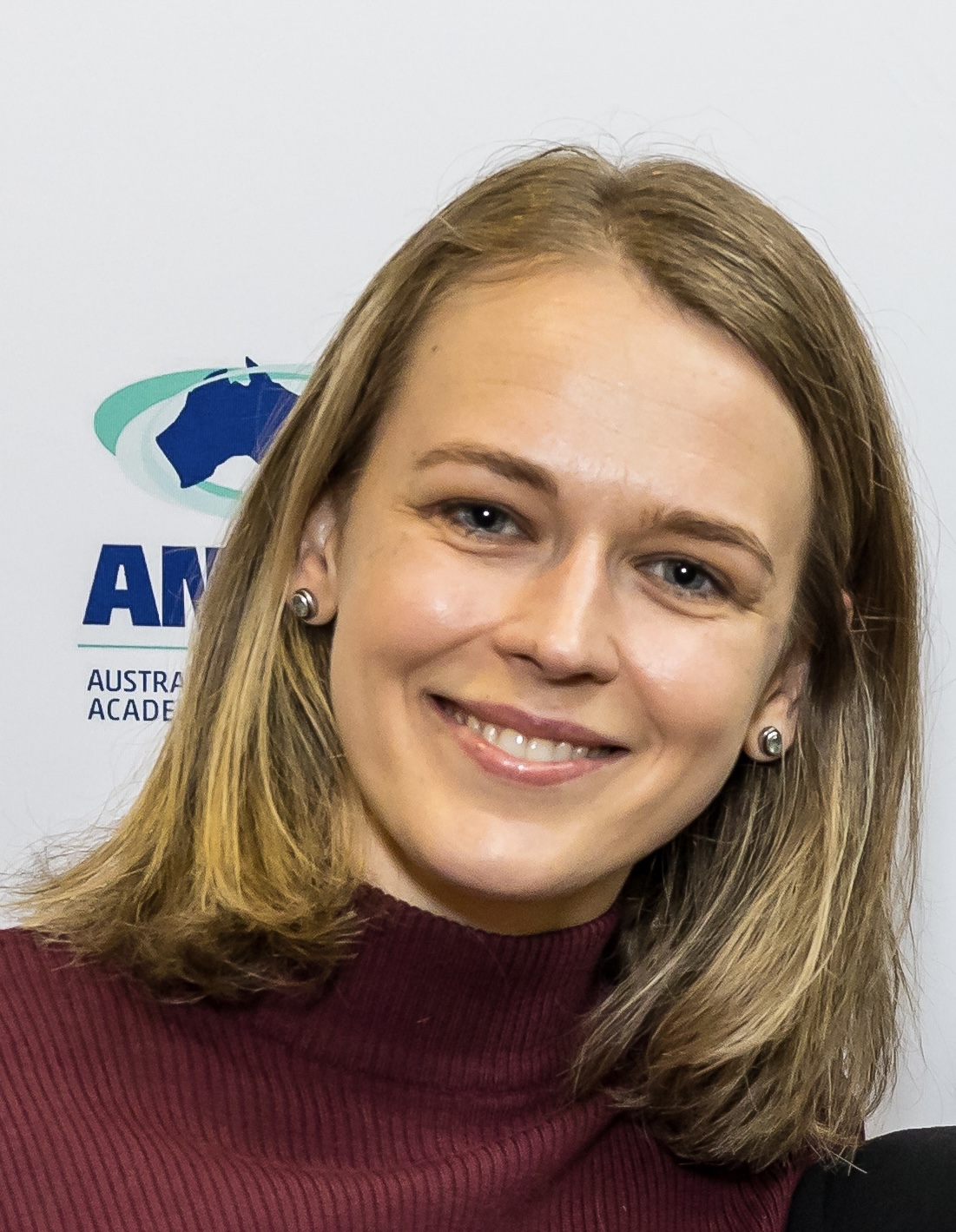 A close-up colour portrait of a smiling woman with straight, shoulder-length blonde hair, wearing stud earrings and a ribbed maroon turtleneck top, standing in front of a white backdrop with a blue and green logo partially visible on the left.