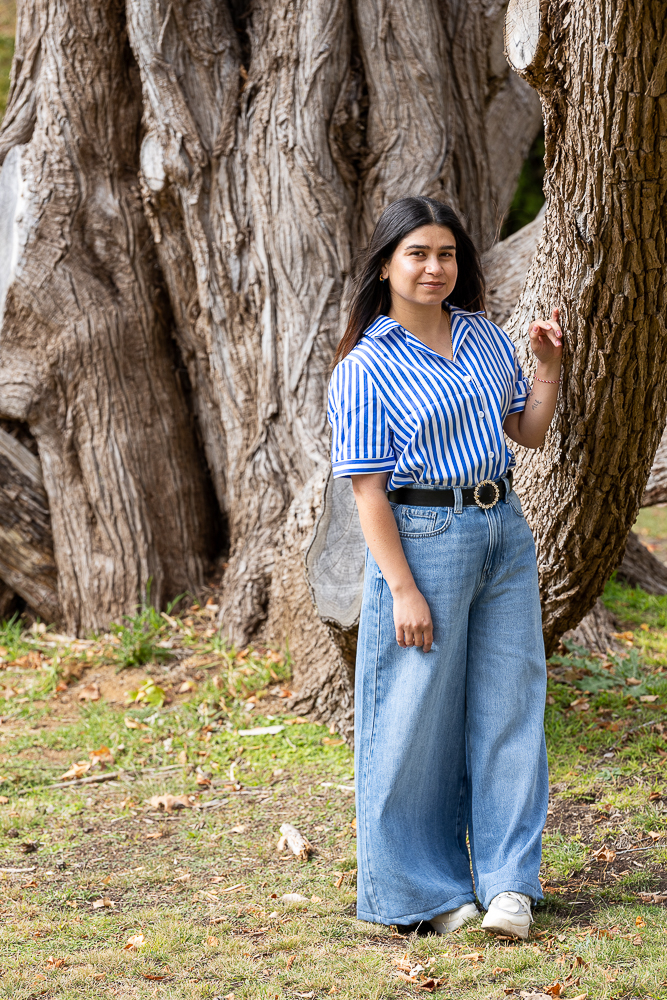 Amera Ali, a young Yazidi woman with long dark hair, stands in front of a huge, textured tree trunk on a grassy area at UNE. She wears a blue‑and‑white vertical striped short‑sleeve shirt tucked into high‑waisted wide‑leg light blue jeans with a black belt, and white sneakers. One hand rests lightly on the tree and she looks towards the camera with a calm, steady expression.