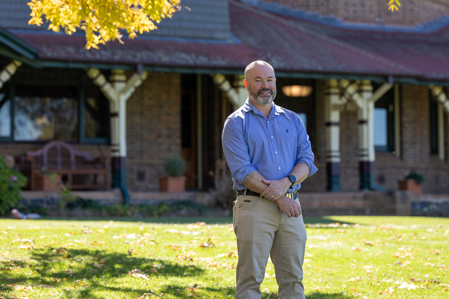 Dane O’Connor stands on the lawn in front of a historic brick homestead at UNE on a sunny autumn day, wearing a light blue shirt and beige pants, smiling with arms loosely folded as golden leaves frame the top of the image.