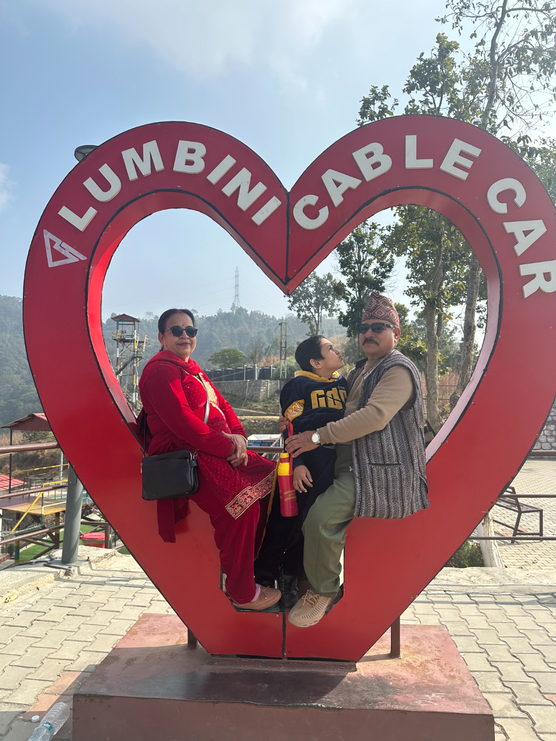 Shila Pokhrel’s parents and young son pose together inside a large red heart-shaped Lumbini Cable Car frame at an outdoor lookout in Nepal. Her father, wearing a patterned topi hat, vest and glasses, sits at the back with his arm around his grandson, who is dressed in a dark jumper and cap. Her mother, dressed in a bright red traditional outfit with gold embroidery, sits in front, smiling, with a small black shoulder bag resting on her lap. Hills, trees and cable car structures are visible in the hazy background.