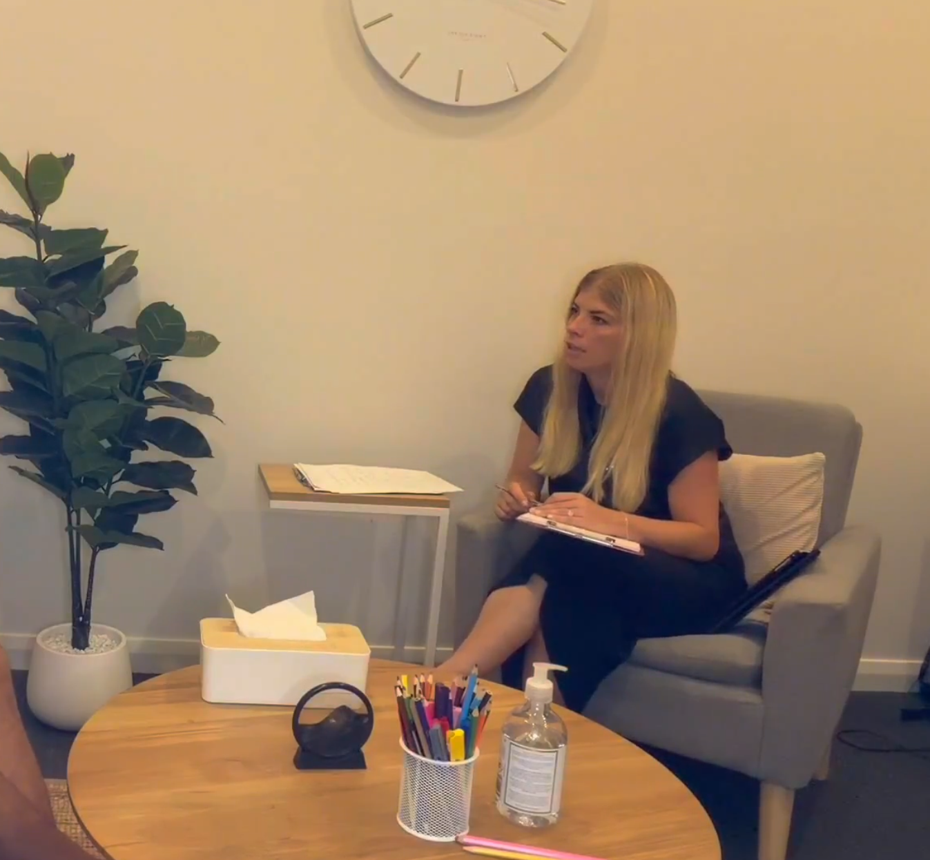 Dr Henri Dohnt sits in a comfortable grey armchair in a calm counselling room, holding a clipboard and pen as she listens attentively to an unseen person opposite her. A small round table with coloured pencils, tissues and hand sanitiser sits in the foreground, with a leafy indoor plant and a large wall clock completing the warm, professional setting.