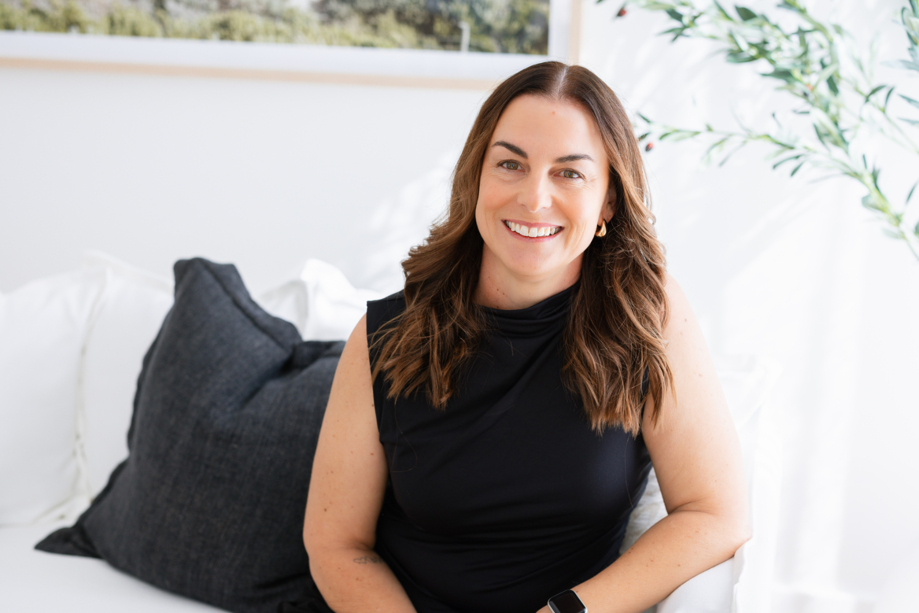 A professional portrait of Olivia Tierney, a woman with long brown hair, smiling warmly at the camera. She is wearing a black sleeveless top and is seated in a bright, light-filled room with soft furnishings and greenery visible in the background. A green 