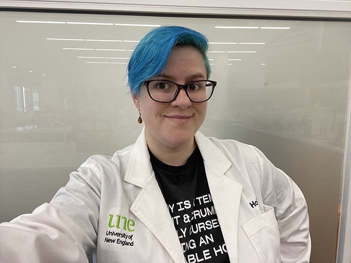 Image of Dr Holland Dougherty smiling at the camera wearing a lab coat. She has blue hair and is wearing glasses.