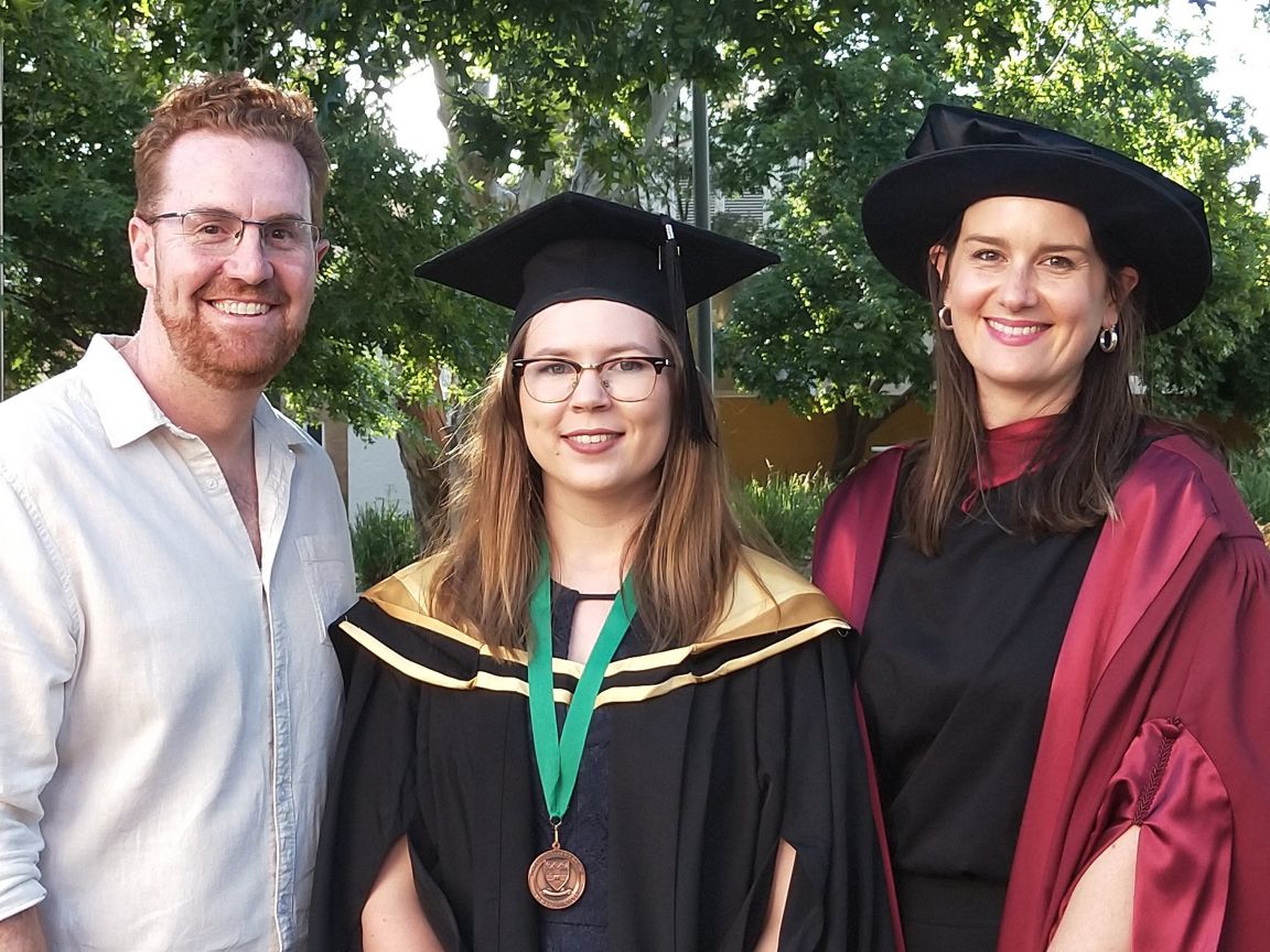 A man and two women stand together, smiling. The man wears a light, collared shirt. The woman in the middle wears a Bachelor’s mortarboard and gown, as well as a medal around her neck on a green ribbon. The second woman wears PhD robes and bonnet.