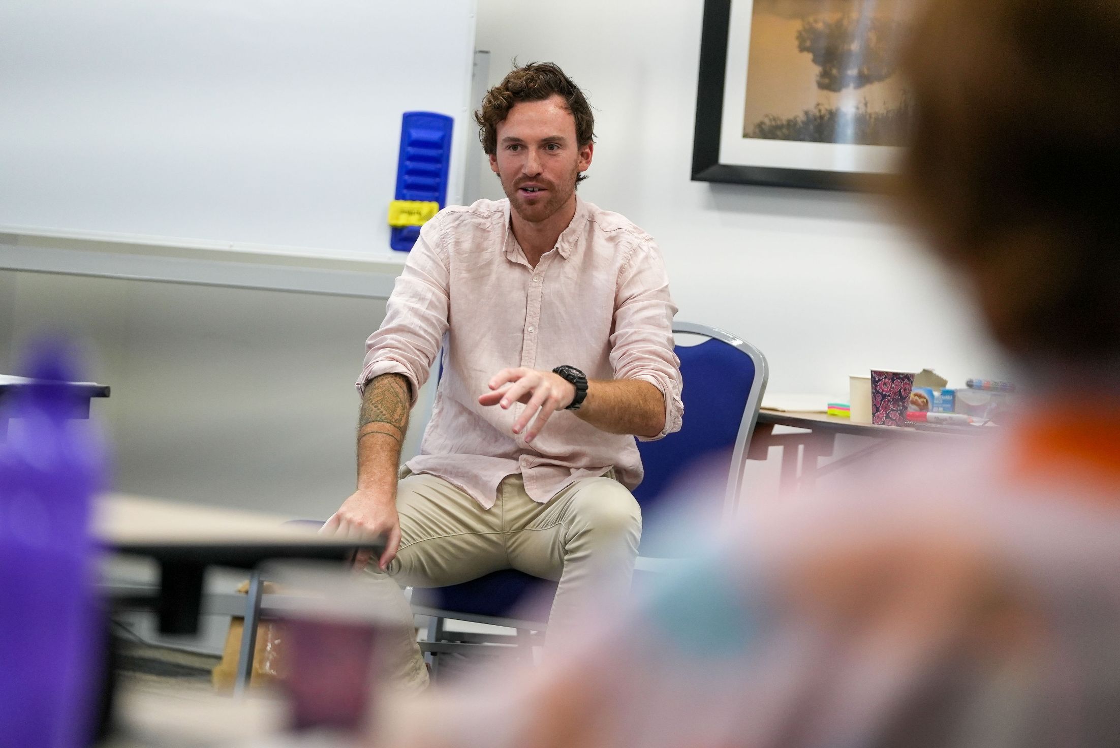 A man with dark hair and a short beard sits in an office environment. He is facing people and is speaking and gesturing. He wears a very pale pink linen shirt and khaki pants and a chunky black watch.