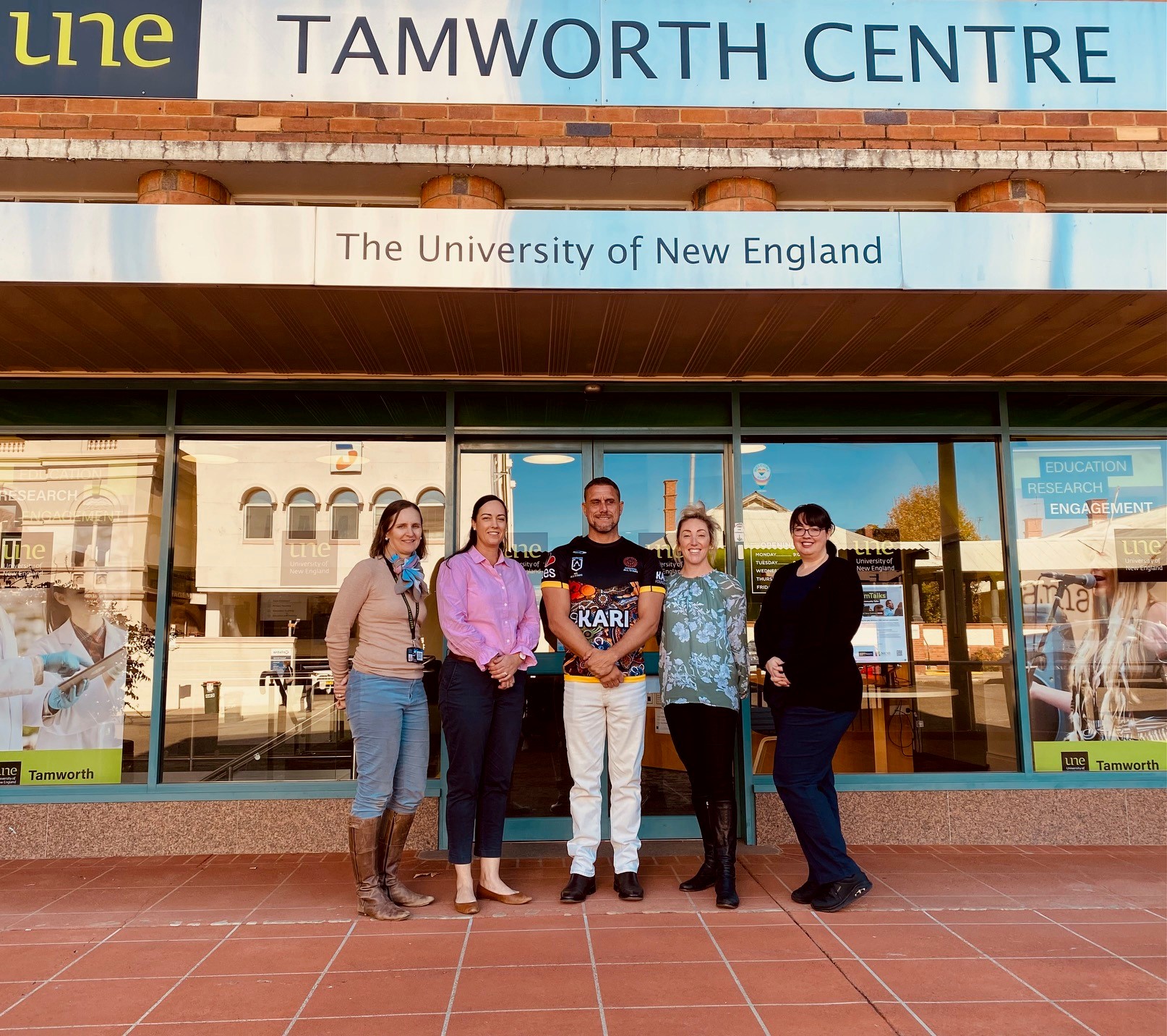 Five smiling UNE Tamworth Staff members stand by the front door of the UNE Tamworth building.