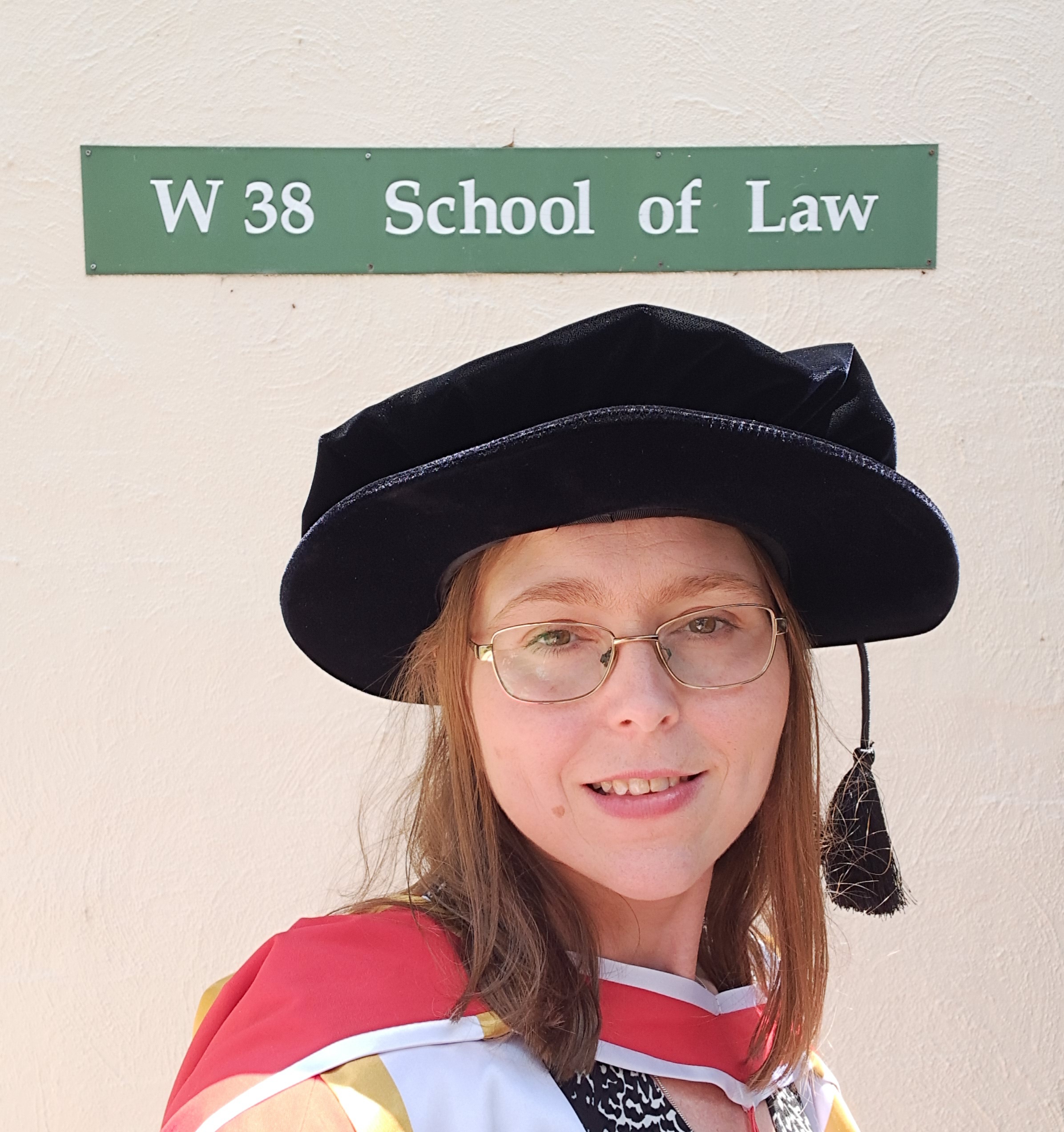 Dr Wellett Potter under Law School sign on campus dressed in formal graduation gown.