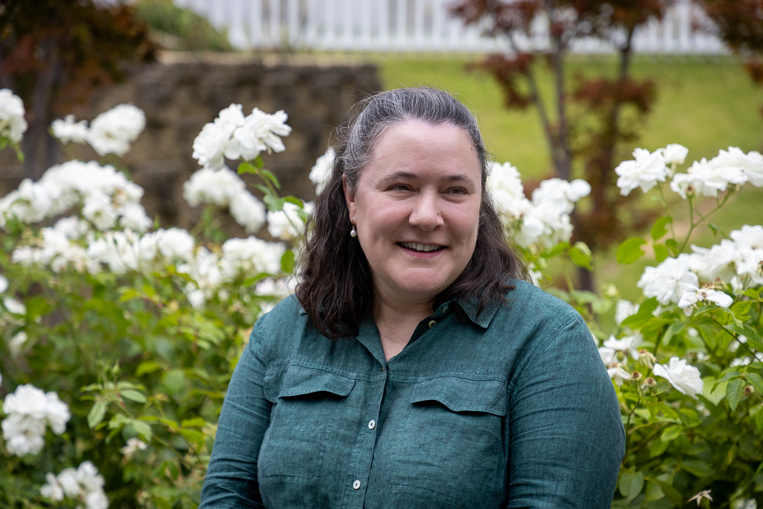 Smiling woman with greying dark hair, wearing a green shirt, sits in front of a garden bed of white roses.