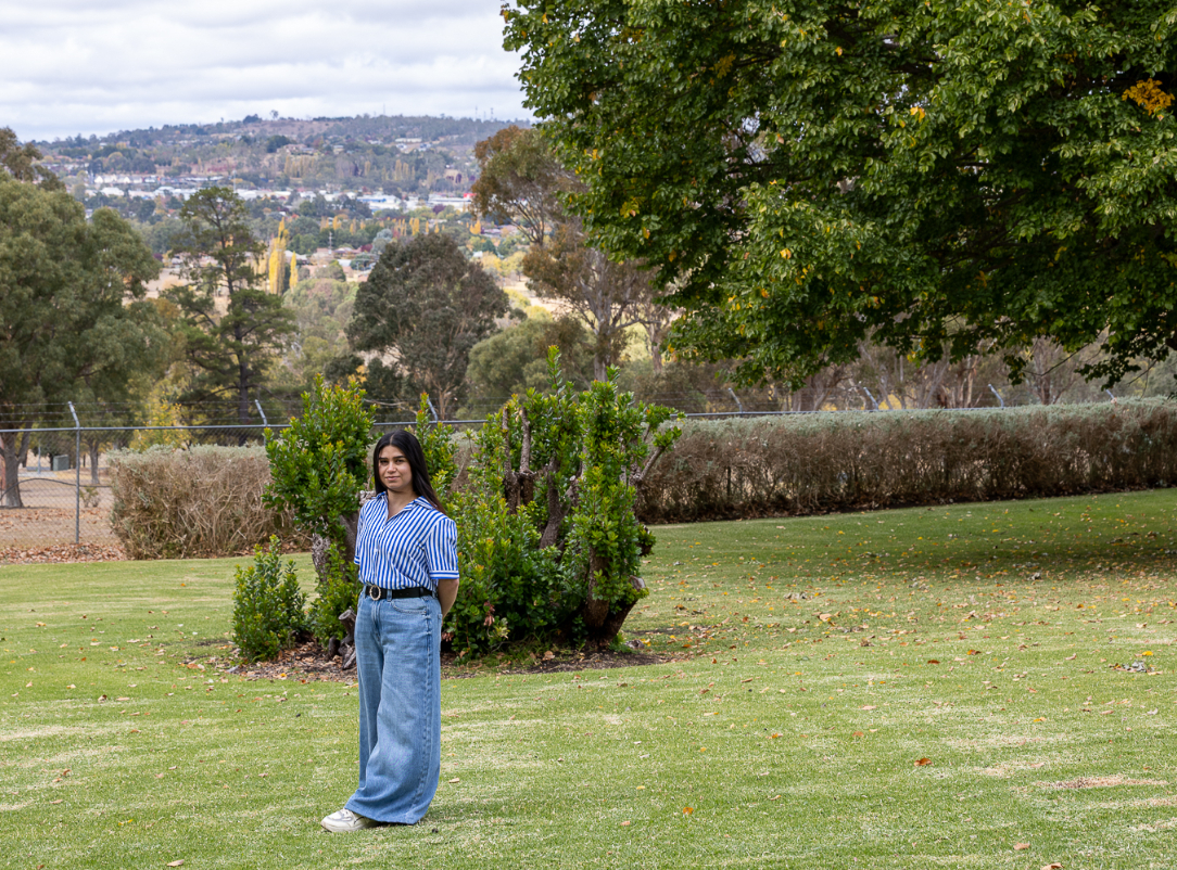 Young woman standing on a large green lawn with her hands behind her back, wearing a blue‑and‑white striped short‑sleeve shirt, wide‑leg denim jeans, and white sneakers. She is positioned near a low, leafy shrub with tall trees and hedges behind her. In the distance, a regional town and rolling hills are visible under an overcast sky, creating a spacious, park‑like campus setting.