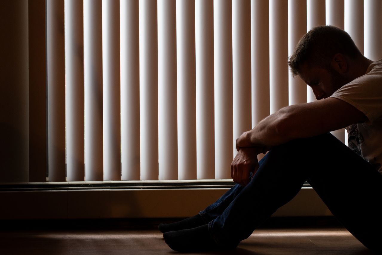 Man with depression sitting next to a window with their head in their hands.