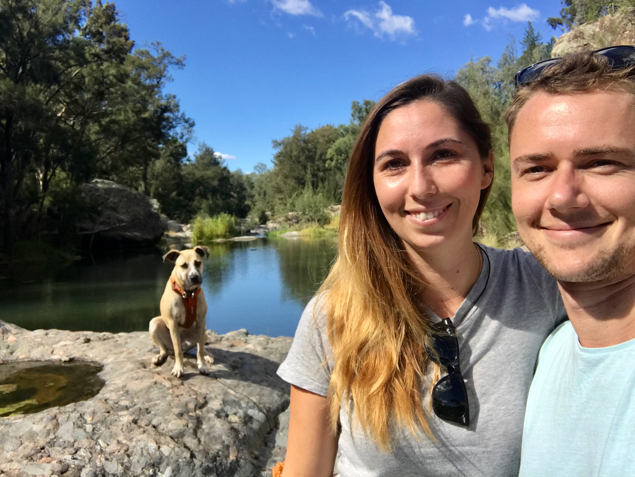 Two adults smile for a selfie beside a calm rocky creek or waterhole, with a tan dog sitting on a rock near the water and bushland trees in the background under a bright blue sky.