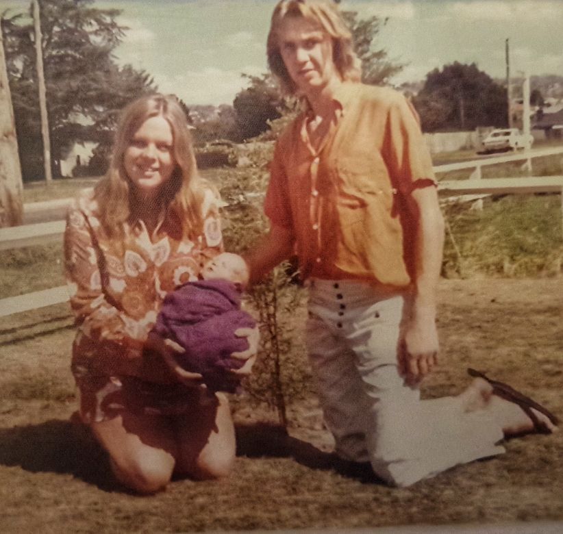 A young man and woman pose on either side of a newly planted tree. The woman has medium-length blonde hair and wears a paisley patterned dress. The man has shoulder-length blond hair, and wears an orange shirt, white pants and dark thongs. The woman holds a small baby wrapped in a purple rug. In the background are low, white wooden fences and an older-model white car.