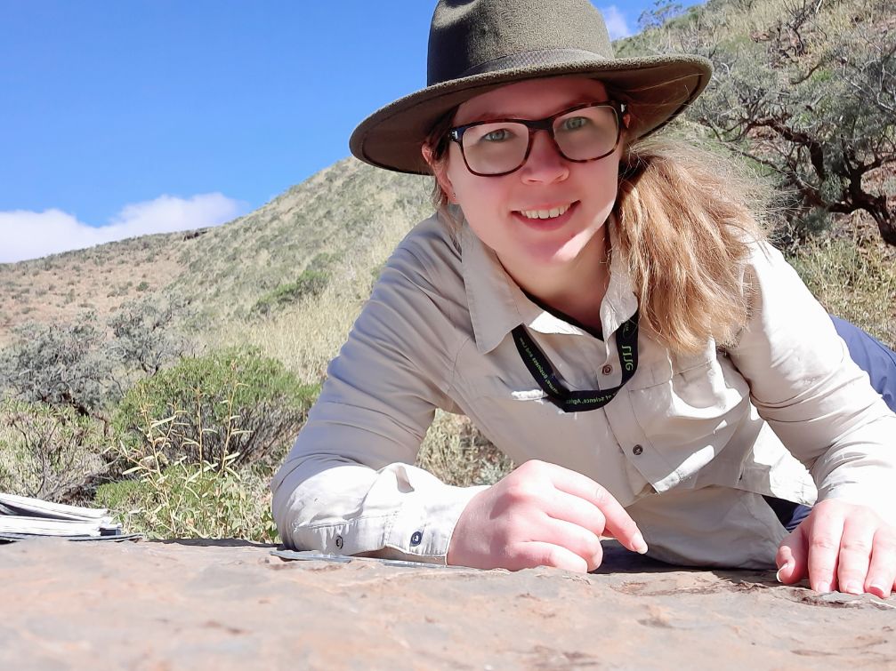 A woman in a broad-brimmed hat lies on some limestone in the bush. She is wearing glasses and leans close to the ground.