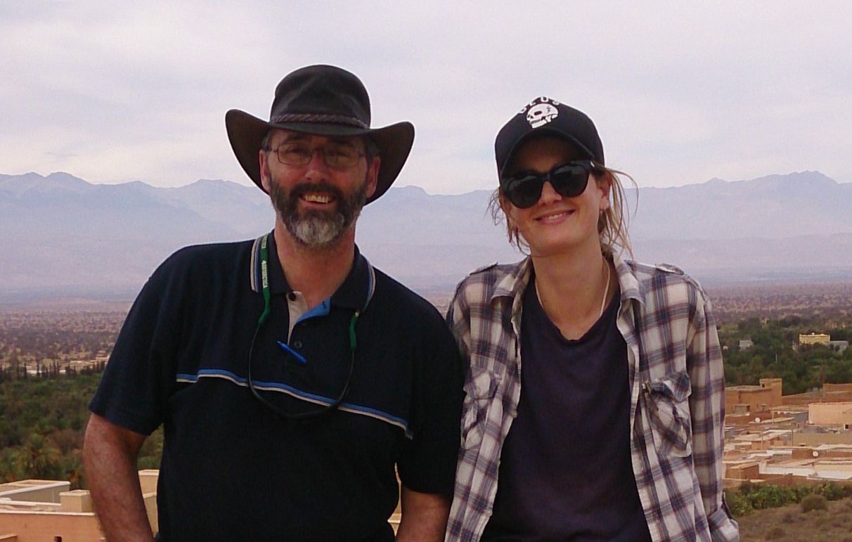 A woman and man stand outdoors, with mountains and buildings in the background. They wear outdoorsy clothing, hats and sunglasses.
