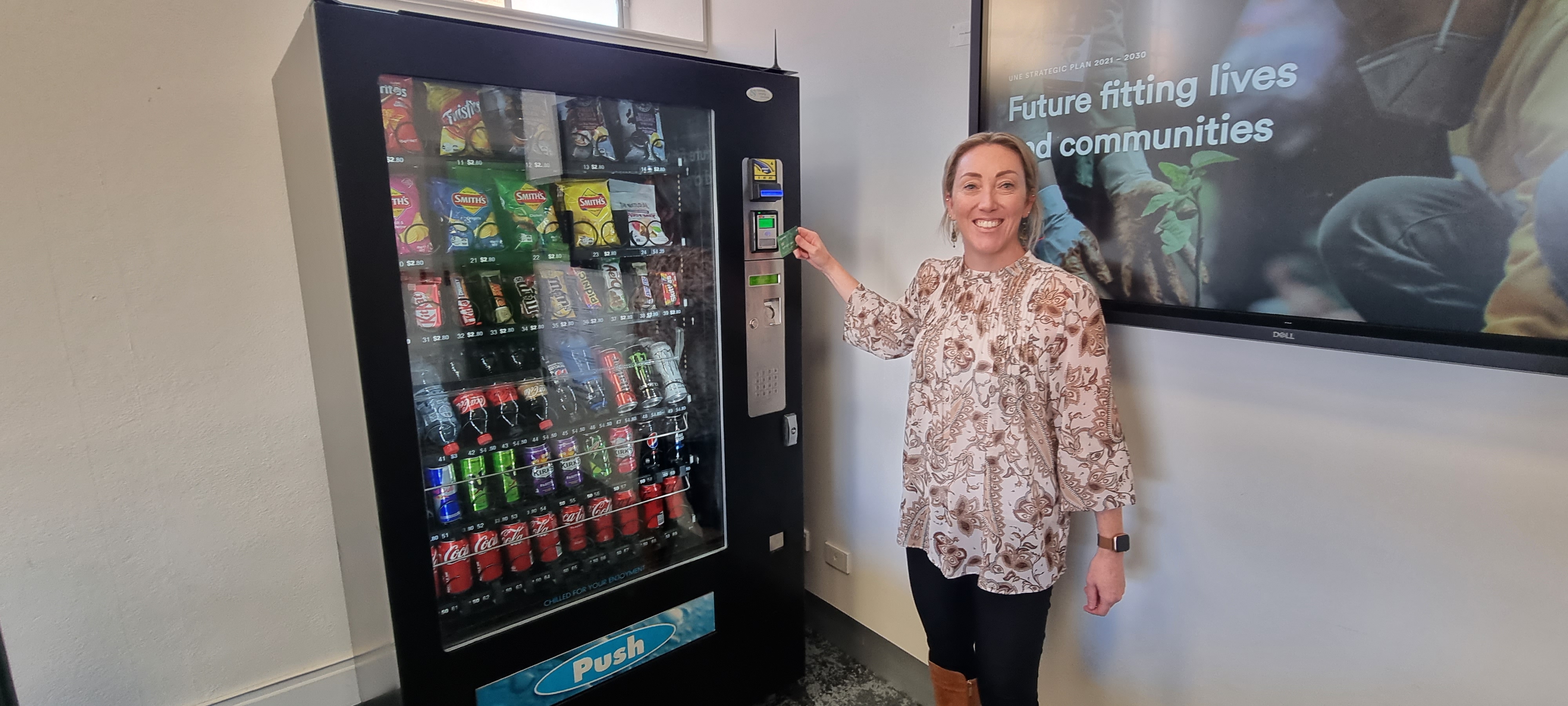 A woman stands before a vending machine, smiling an invitation to purchase some tasty treats