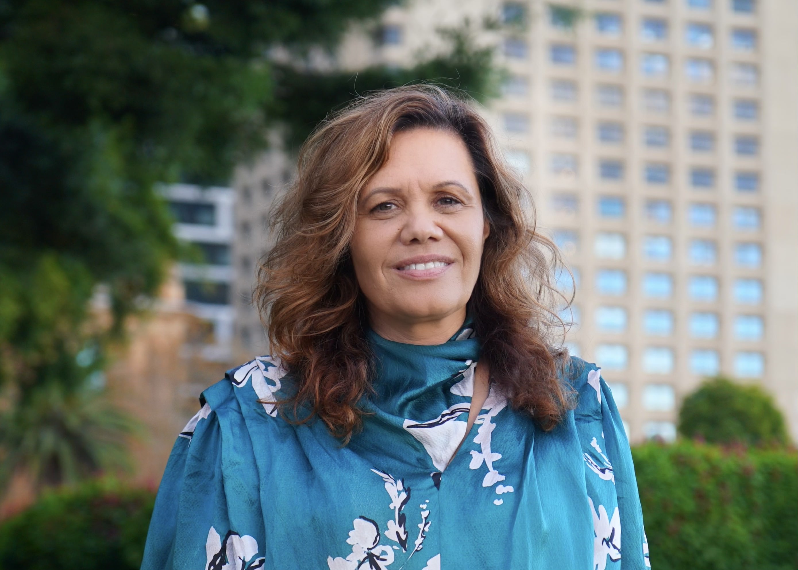 An Indigenous woman with wavy brown hair, wearing a teal top with a white floral pattern, stands outdoors in front of tall buildings and greenery, smiling warmly at the camera.