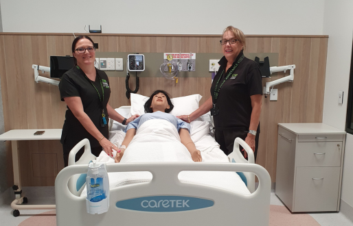 Two UNE nursing educators in black clinical uniforms stand on either side of a hospital bed in of a hospital bed in UNE Sydney nursing simulation lab.