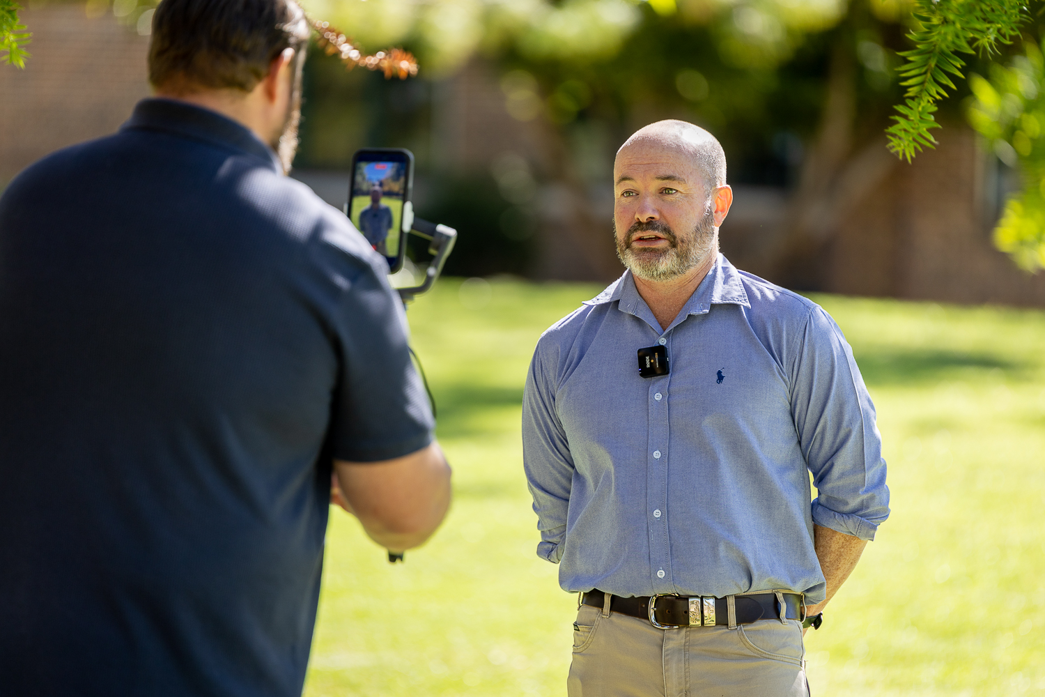 Dane O’Connor stands outside on a sunny day at UNE, wearing a light blue shirt with a microphone clipped to his chest, speaking to an interviewer who films him on a smartphone mounted on a gimbal.
