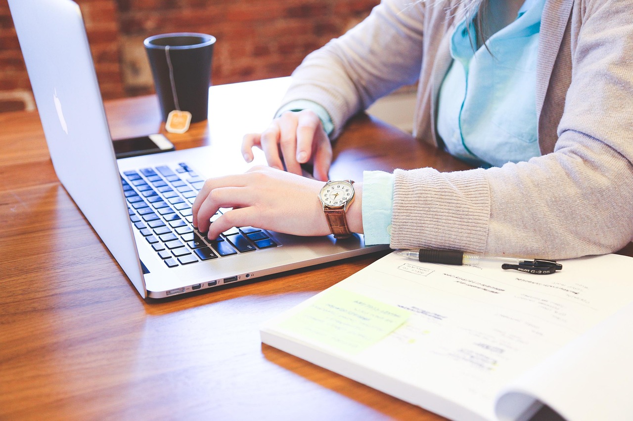 A student sits at an open laptop, with a cup of tea, books, and their glasses next to them on the desk. The student is only shown from the shoulders down. They are wearing a watch.