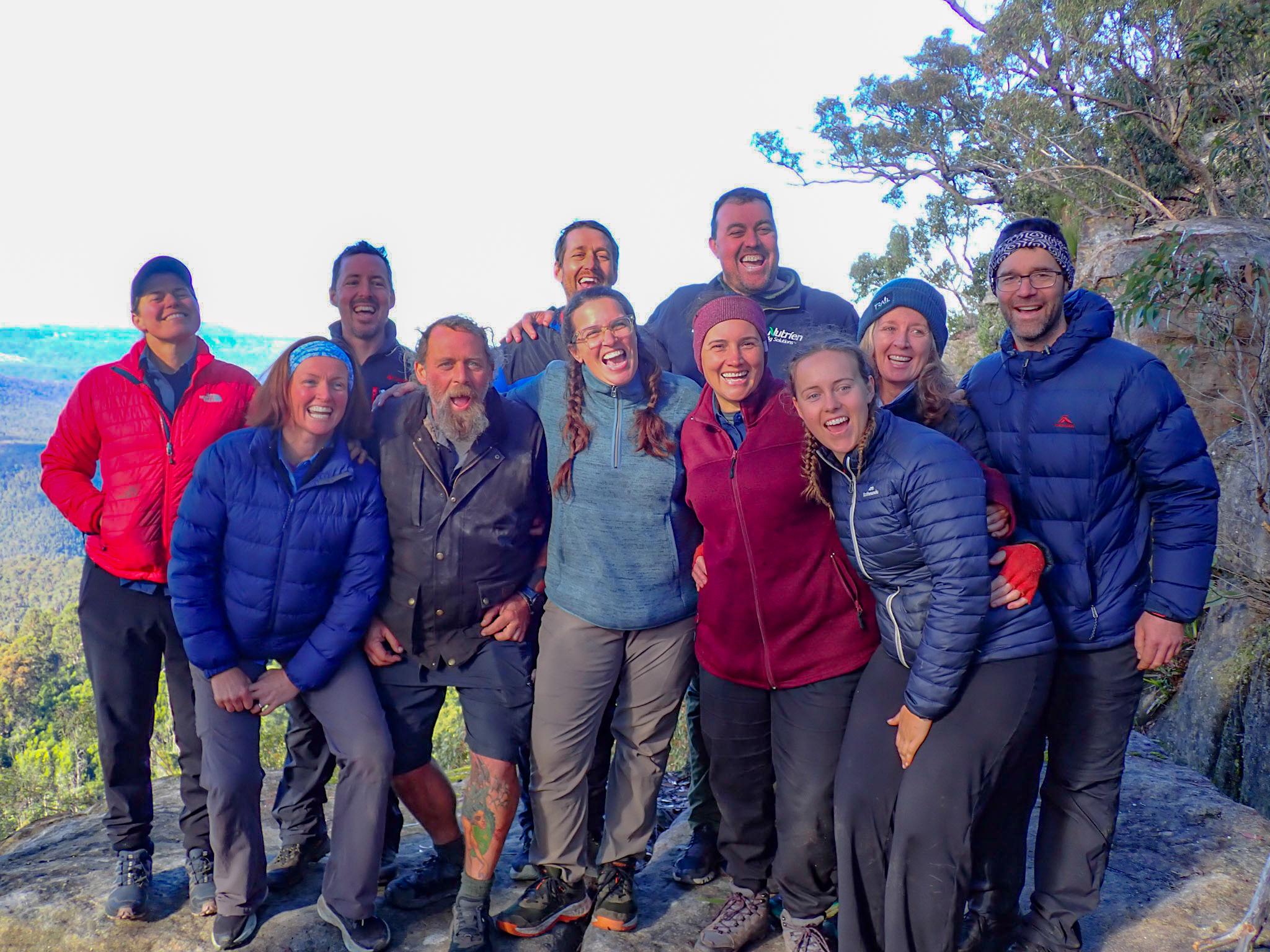 Eleven people in warm outdoor jackets smile together on a rocky lookout, with bushland and a wide valley in the background.
