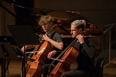 Two male students playing cellos with music stands on stage