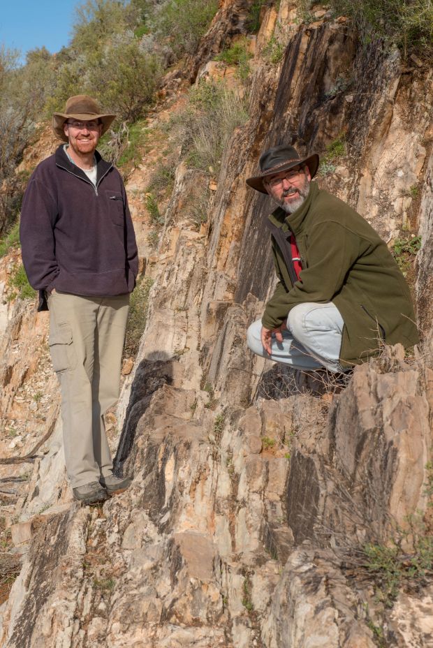 Two men stand on a rocky outcrop in the bush. They both have beards and are wearing hats and outdoorsy clothing. 