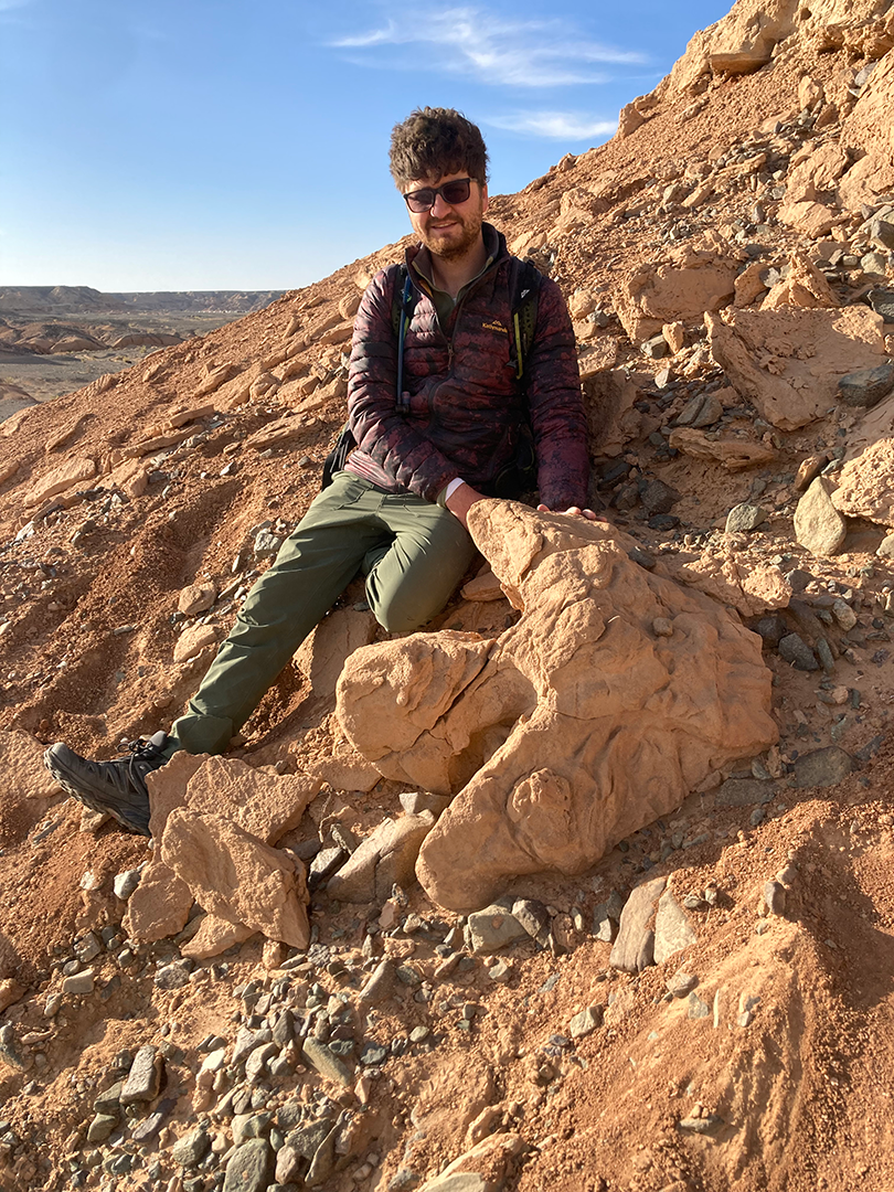 Nathan Enriquez sitting next to a large three-toed hadrosaurid footprint 