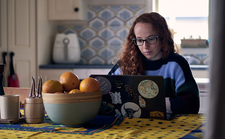 Student with glasses studying on laptop at home kitchen table, with bowl of oranges and blue patterned decor visible