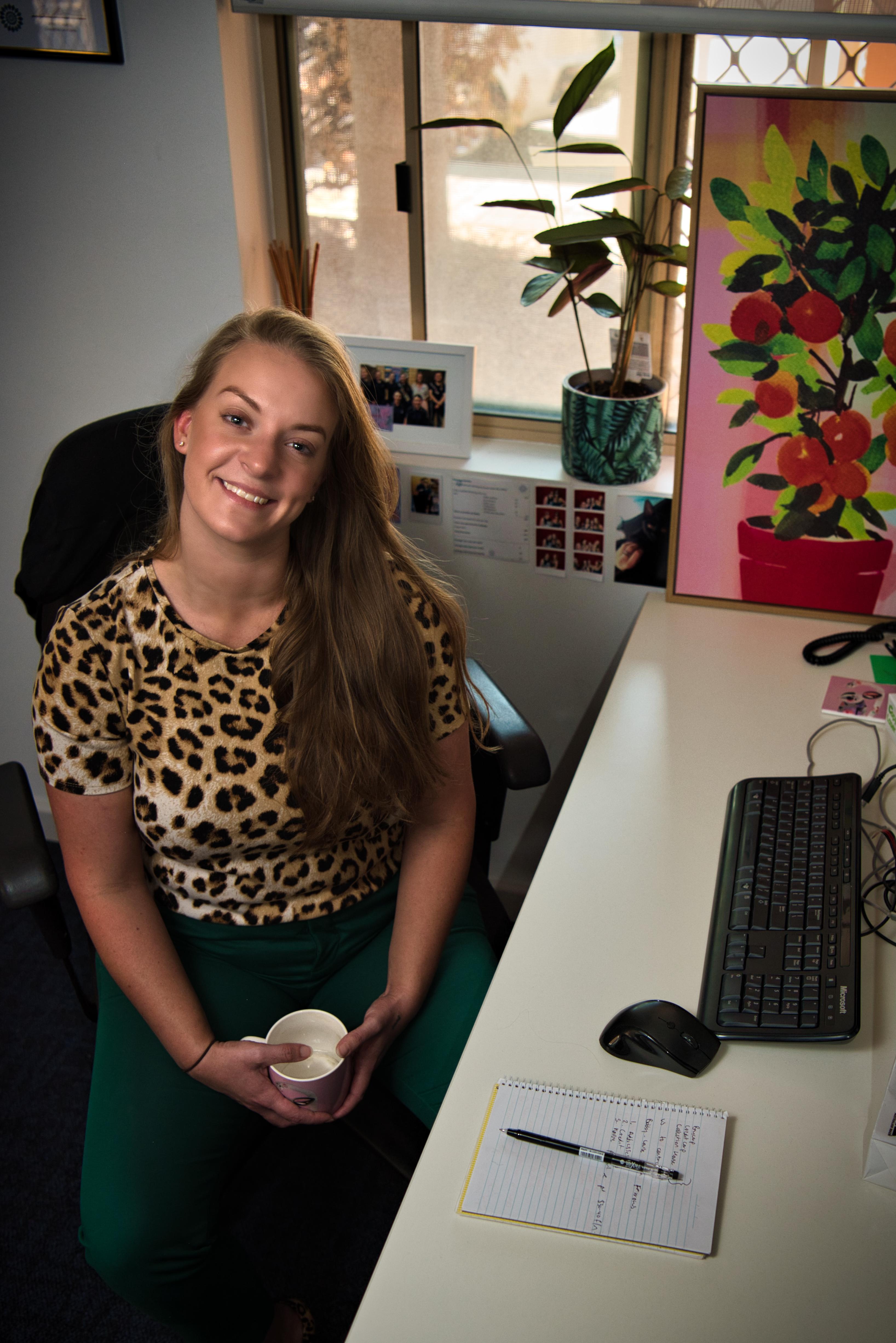 Young woman sitting at an office desk, smiling up at the camera with a mug in her hands.