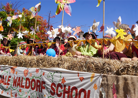 Waldorf School float in the Armidale Autumn Festival parade, featuring children seated among oversized colourful flowers and pinwheels in autumn hues of red, yellow, and white against a bright blue sky