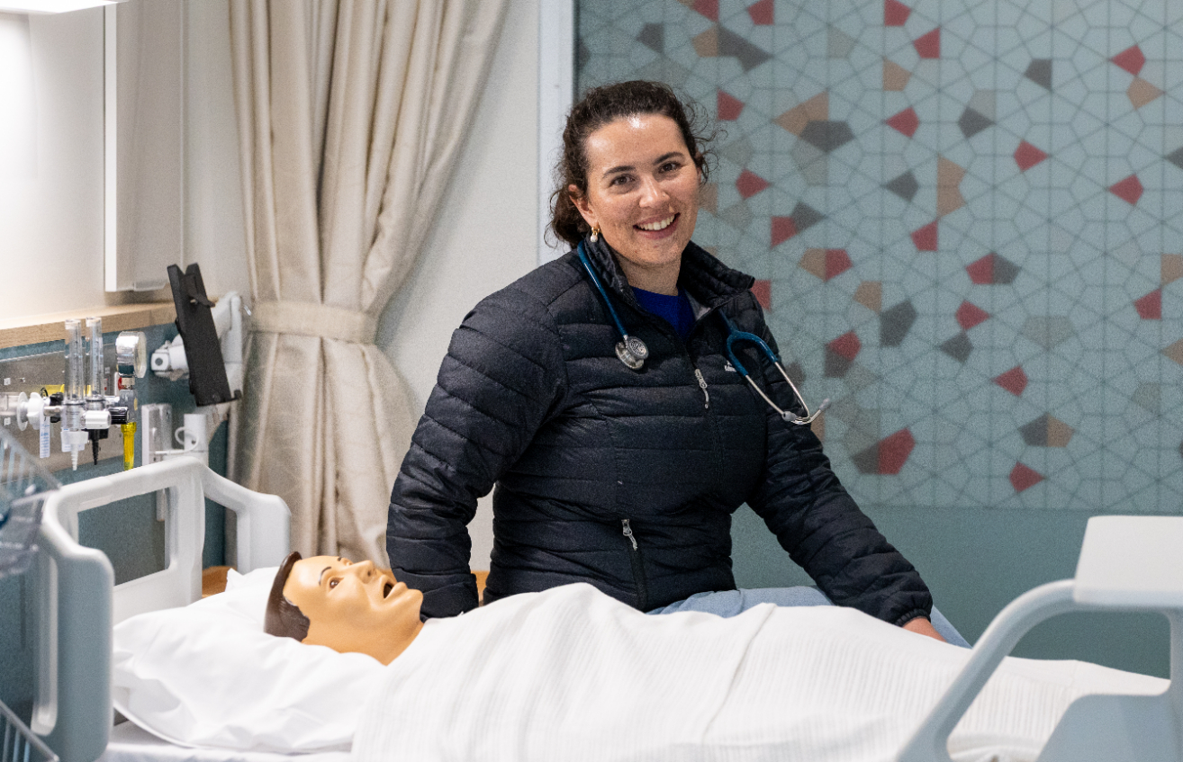 Smiling medical student wearing a dark puffer jacket and stethoscope sits on the edge of a hospital bed in a clinical skills lab, with a lifelike training mannequin lying under a white blanket in the foreground, medical equipment on the wall to the left, and a patterned glass partition in the background.