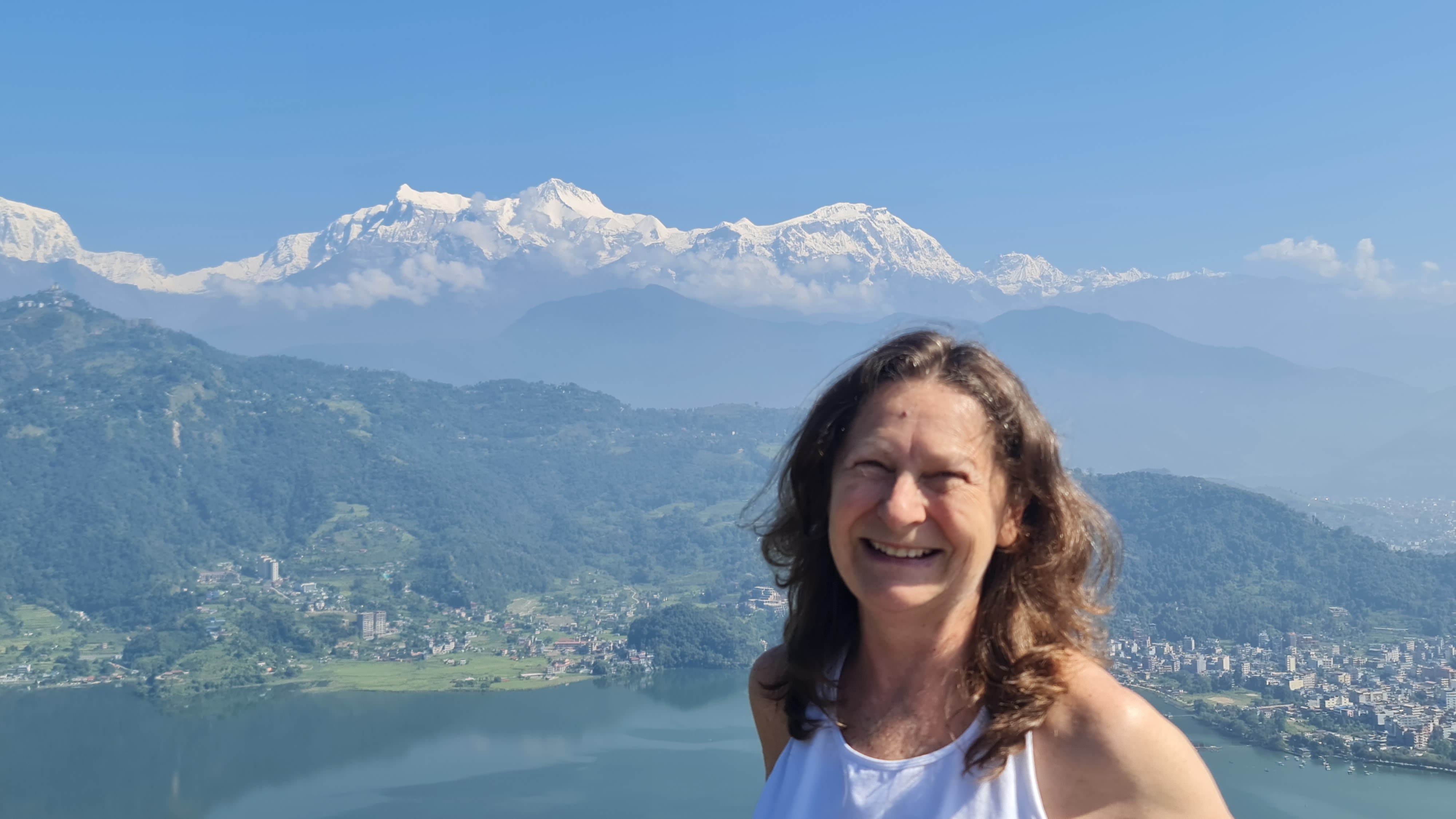 Associate Professor Susan Watt smiling outdoors in a close-up photo, wearing a white sleeveless top, with a lake and town in a green valley behind her and snow-capped mountains under a clear blue sky in the distance.