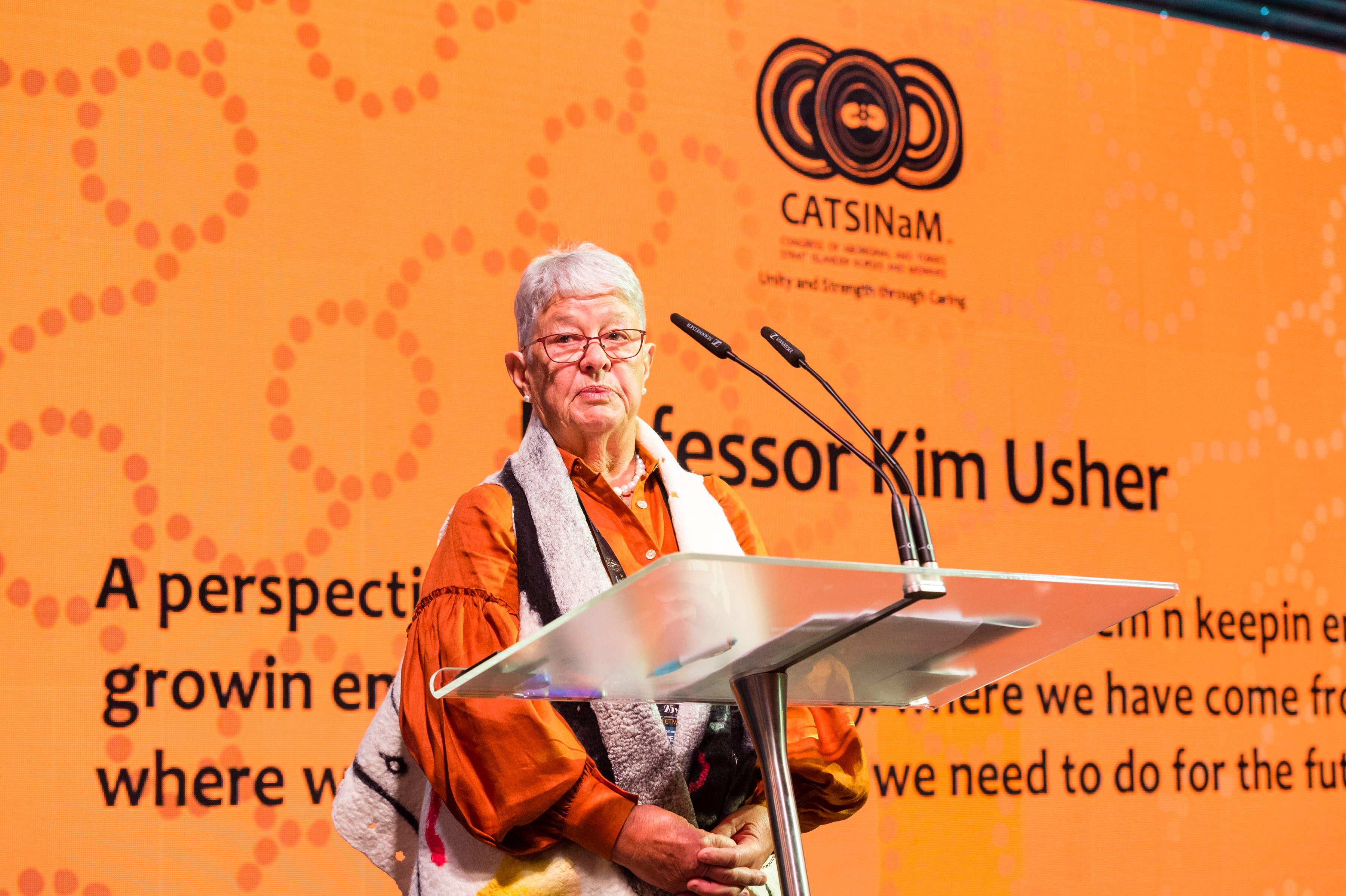 Senior woman wearing orange shirt on stage at podium looks at camera