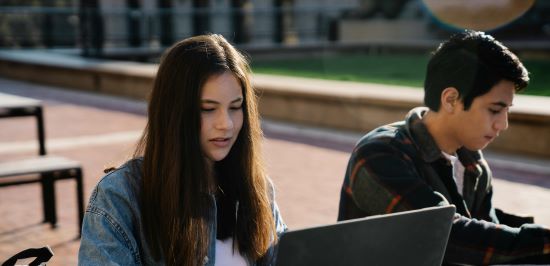 A young female student sitting outside at a table with her laptop open on the desk and a male friend sitting next to her in the background. 