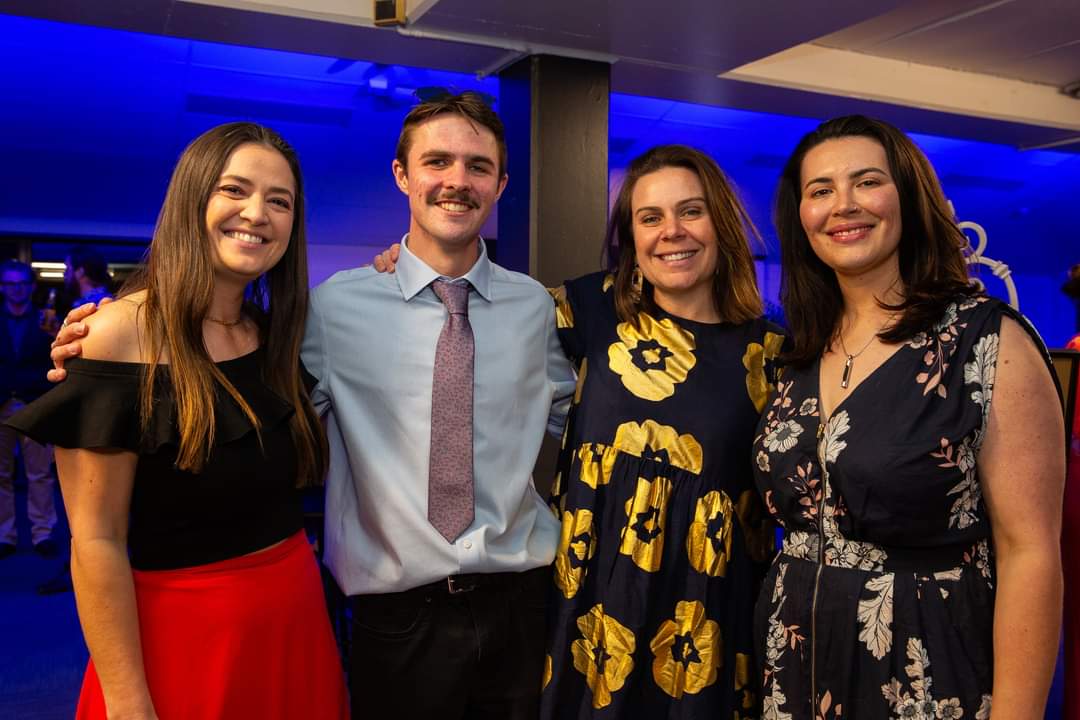 Four young adults stand close together and smile at the camera at an indoor event with blue lighting in the background. From left to right: a woman in a black off‑the‑shoulder top and bright red skirt, a man in a light blue shirt and patterned tie, a woman in a navy dress with large yellow floral prints, and a woman in a dark floral dress.