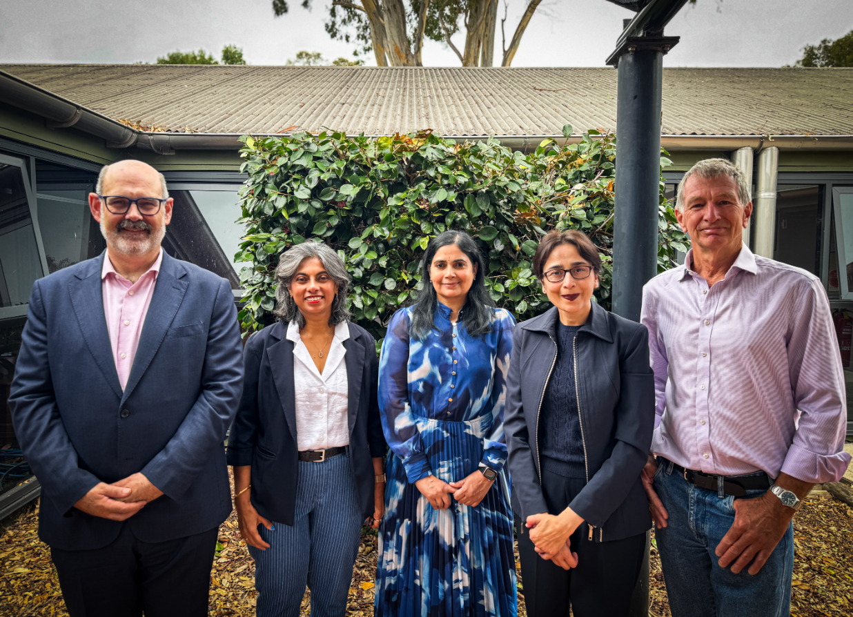 Five adults stand in a row outdoors in front of a green leafy hedge and low building on an overcast day. From left to right: a man in a navy suit and pale pink shirt with glasses and a neatly trimmed beard; a woman with short grey hair wearing a dark blazer, white shirt and blue pinstripe trousers; a woman with long dark hair wearing a blue patterned dress; a woman with dark hair tied back, glasses, and a dark jacket over a navy top and trousers; and a man in a light mauve shirt and blue jeans. They are all facing the camera and smiling softly.