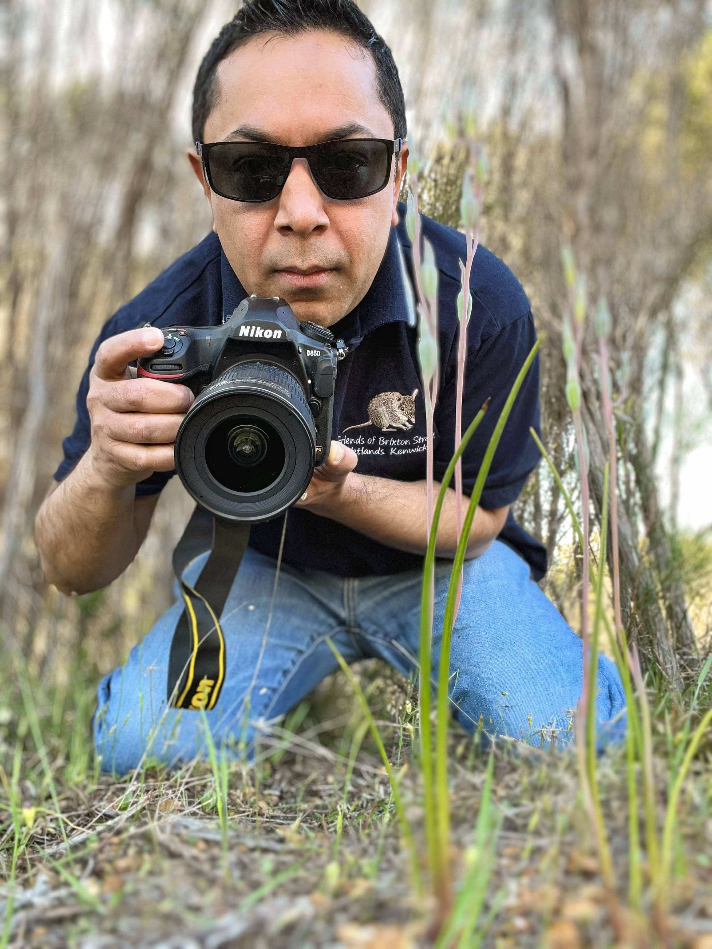 A man wearing dark sunglasses and a navy polo shirt kneels on the ground in a bushland setting, holding a Nikon DSLR camera close to the ground as if about to photograph something at grass level. Tall blades of grass and plants are in soft focus in the foreground, framing him, while the background is blurred scrub and trees, giving the image a natural, outdoor feel.